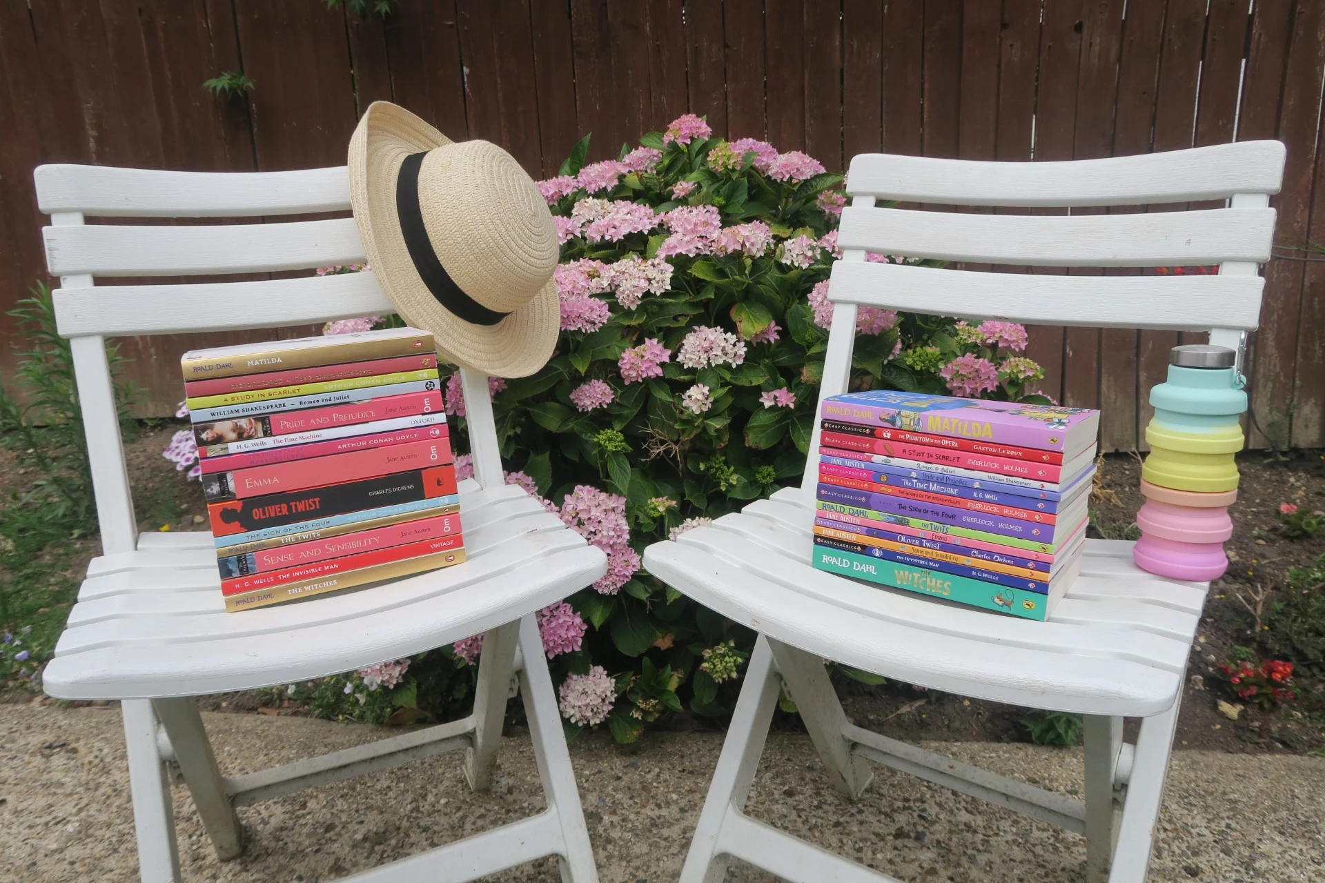 Two Chairs With One Pile Of Adult Books With A Sun Hat And One Deck Chair With The Child Version Of Those Books With A Colourful Water Bottle
