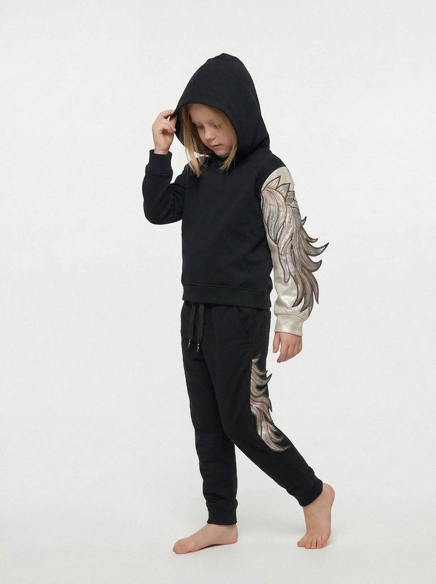 AI generated image with On-Model.com, A child in a black hoodie and pants with metallic silver wing designs on the sleeve and pant leg, standing baref