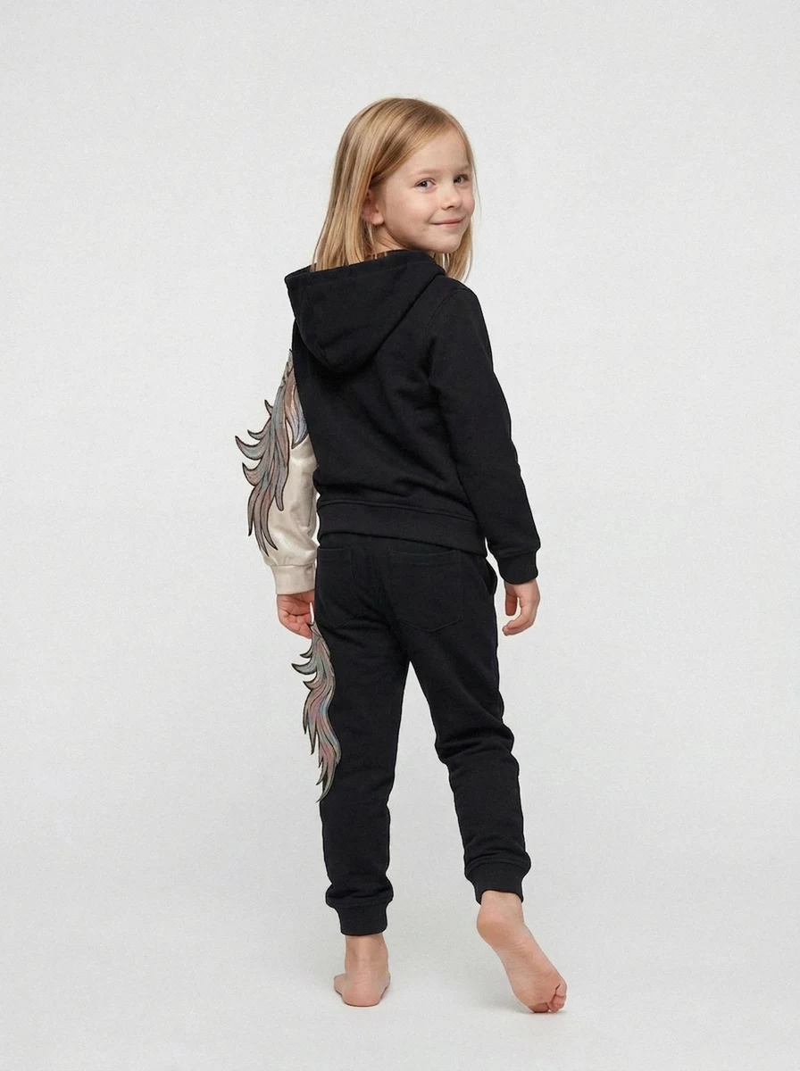 PDP image generated with On-Model.com A smiling young girl in a black hoodie with colorful feather designs on the sleeves and pants, standing barefoot